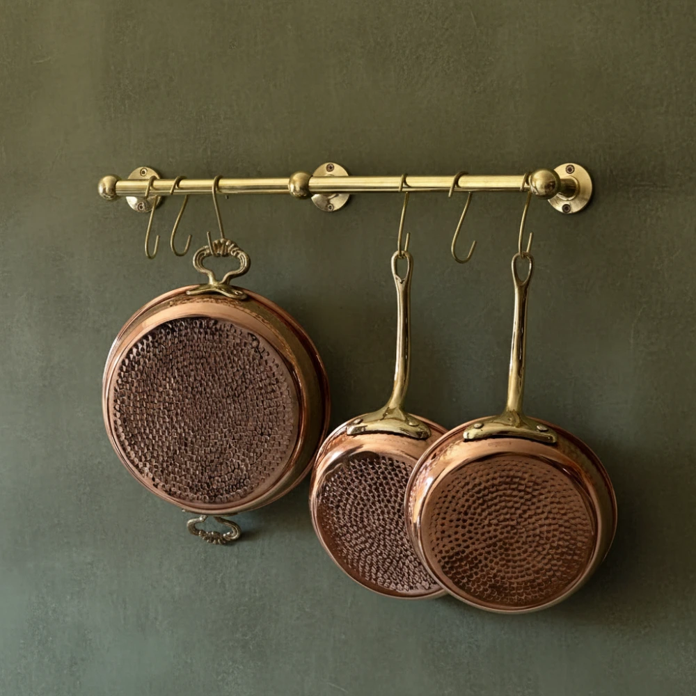 Three hammered copper pans with brass handles hanging from a brass rail against a muted green wall.