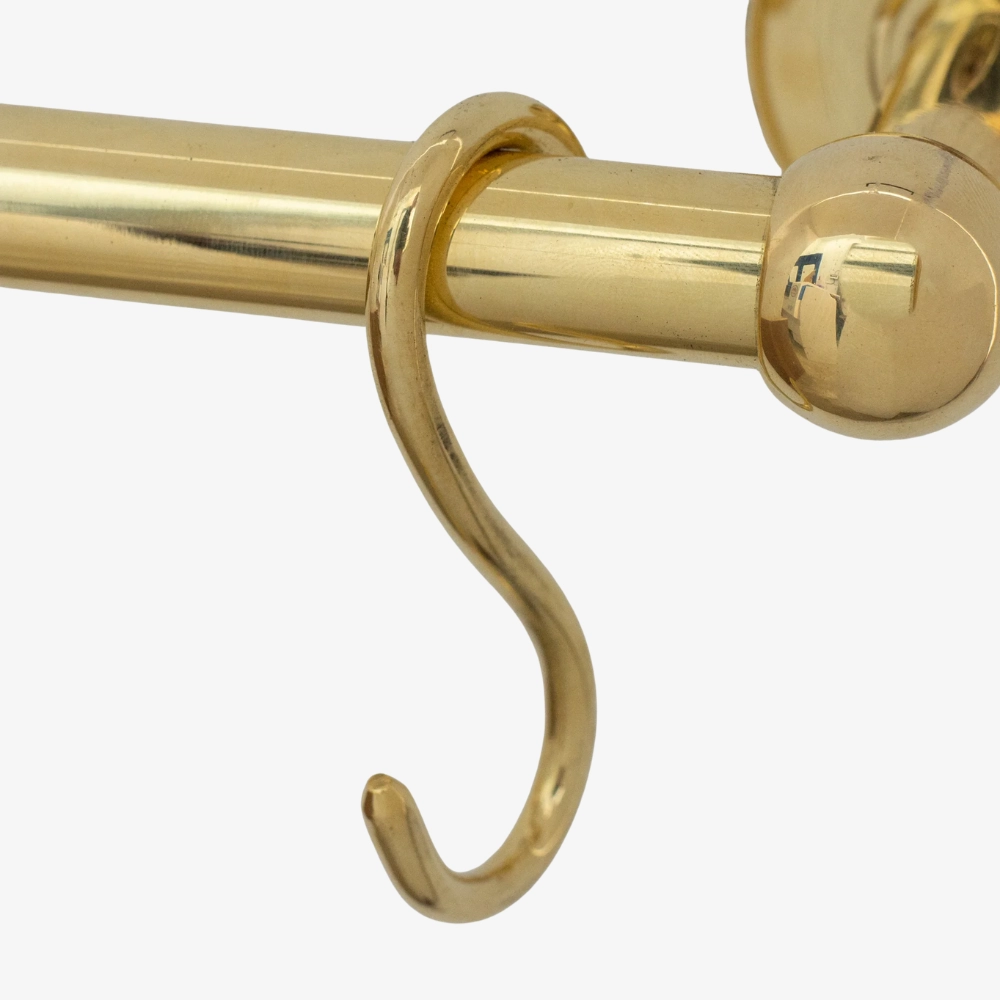 Polished brass S-shaped hook hanging from a brass curtain rod near a rounded finial, close-up.