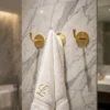 White towel embroidered with gold L hanging on a central brass hook against veined marble, flanked by two empty brass hooks.