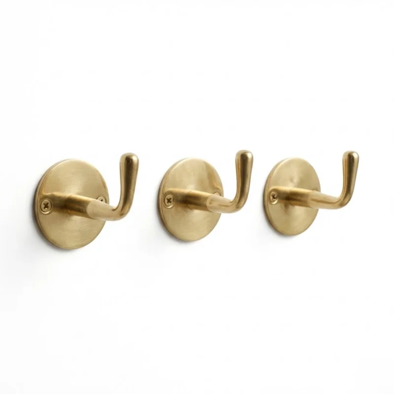 Three brass wall hooks with round bases and single visible screws mounted on a white wall.