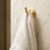 White embroidered towel hanging from a brass wall hook mounted on a light marble wall.