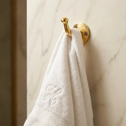 White embroidered towel hanging from a brass wall hook mounted on a light marble wall.