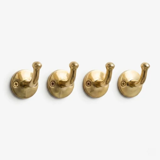 Four polished brass wall hooks with round backplates and upward-curving pegs mounted in a row on a white wall.