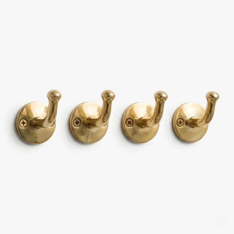 Four polished brass wall hooks with round backplates and upward-curving pegs mounted in a row on a white wall.