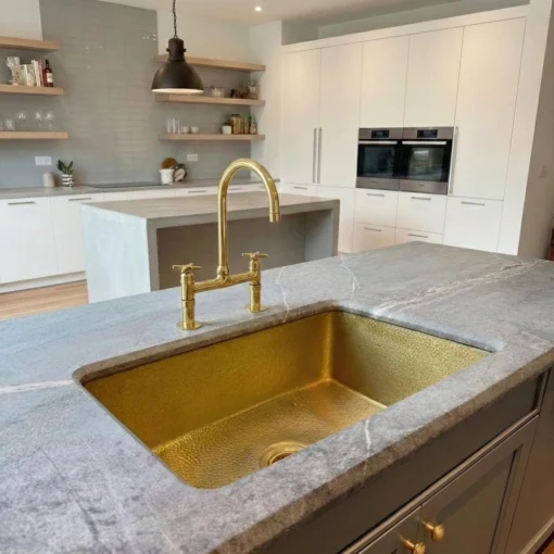 Hammered brass kitchen sink with matching gold bridge faucet set in a gray marble countertop in a modern white kitchen.