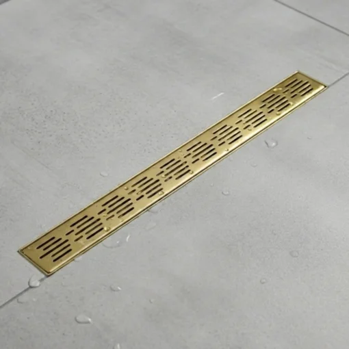 Long brass linear shower drain set in light gray tiled floor with scattered water droplets.