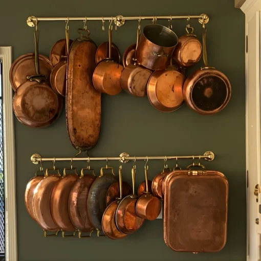 Assorted vintage copper pots, pans, and a rectangular roasting pan hanging on brass rails against a green wall.