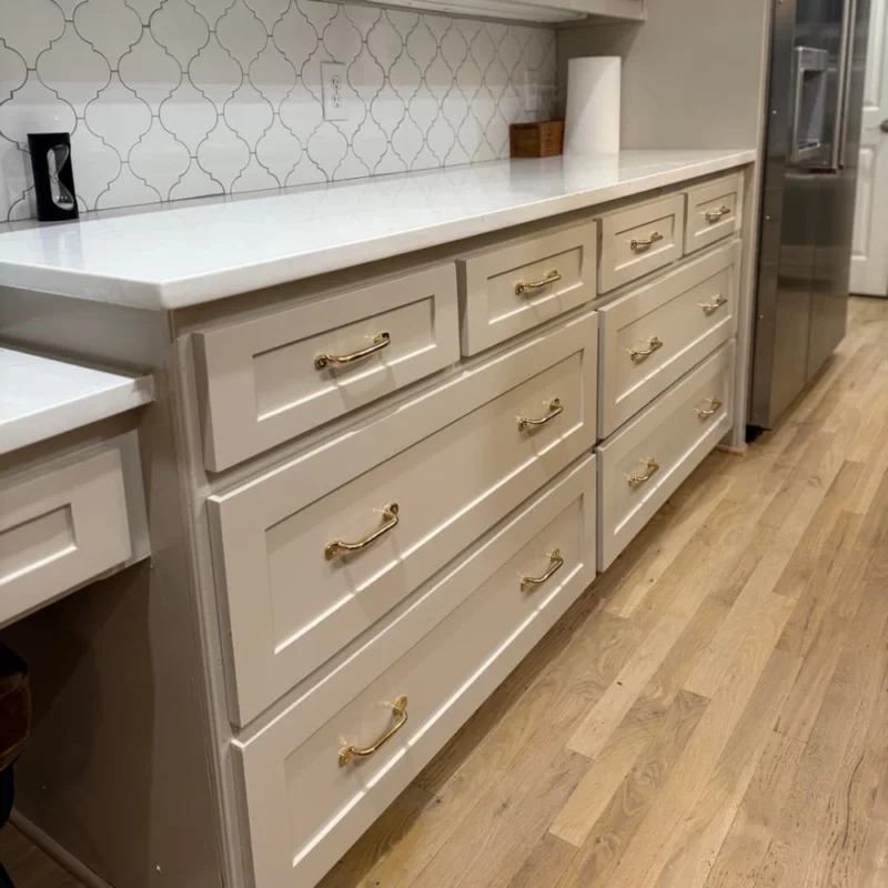 White kitchen drawers with brass handles under a white quartz countertop and patterned tile backsplash on a hardwood floor.