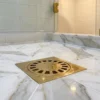 Brass square shower drain with circular grate set in veined white marble tiles, droplets on the surface.