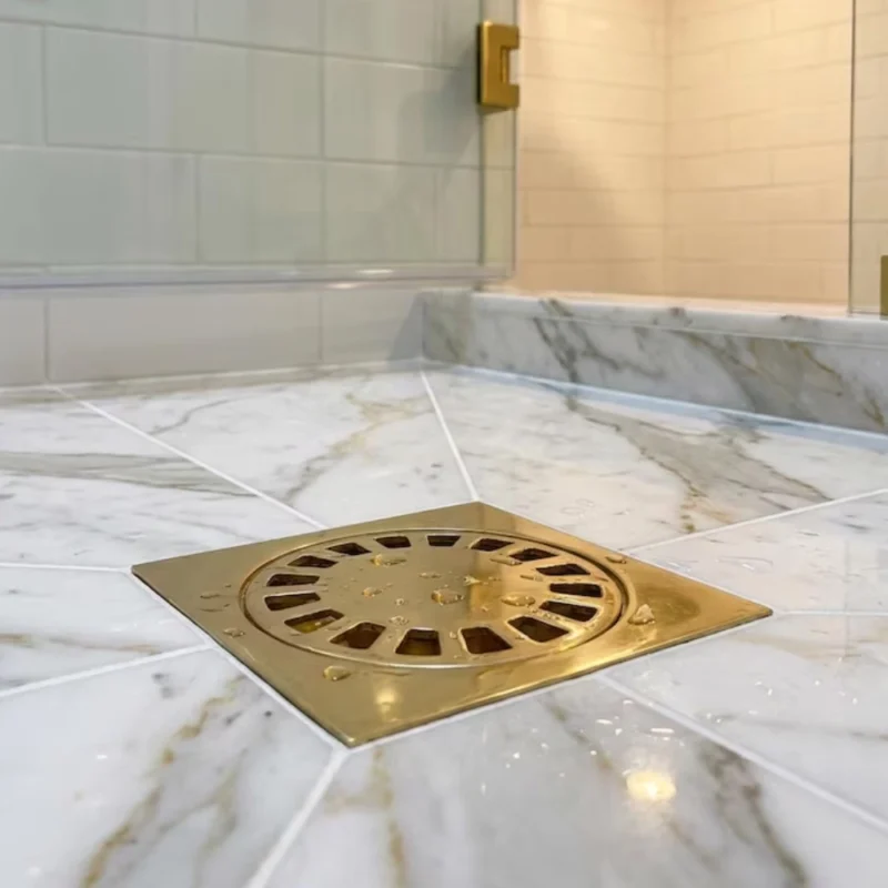 Brass square shower drain with circular grate set in veined white marble tiles, droplets on the surface.