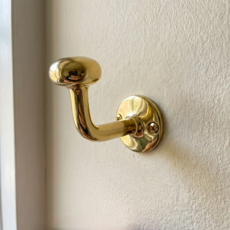 brass coat hook with round base and visible screws mounted on a textured off-white wall, reflecting its surroundings.