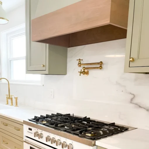 Gas range with black grates beneath a wooden hood, marble backsplash featuring a brass pot filler and sage-green cabinets.