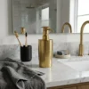 Brass soap dispenser on white marble bathroom counter with matching brass faucet, toothbrushes in black cup, and gray towel.
