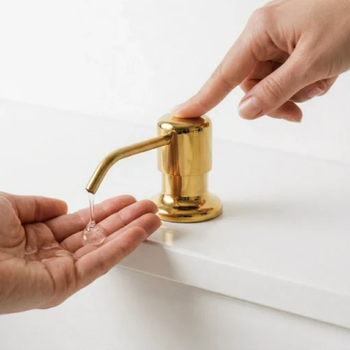 unlacquered brass soap dispenser being pressed to release clear liquid soap into a cupped hand on a white countertop.