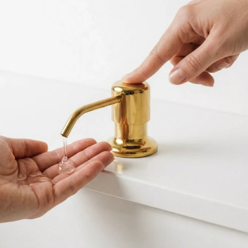 unlacquered brass soap dispenser being pressed to release clear liquid soap into a cupped hand on a white countertop.